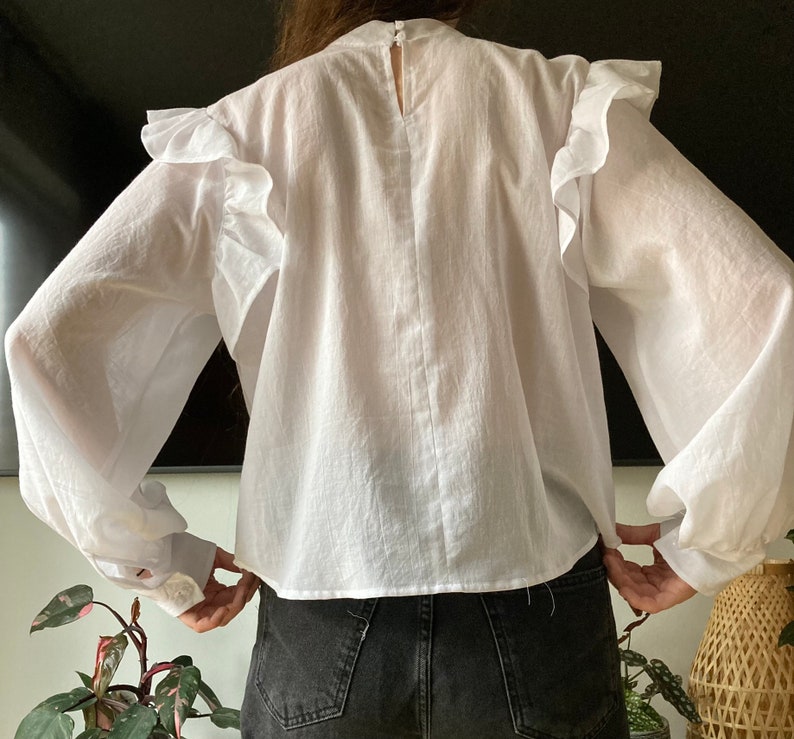 May include: White long-sleeve blouse with a ruffled neckline and puffed sleeves. The blouse has a relaxed fit and a slightly cropped hem.