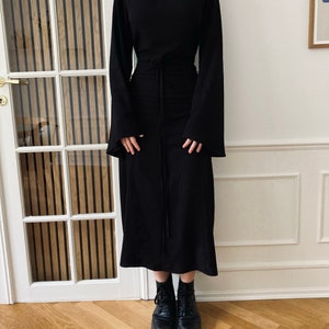 Puede incluir: Un vestido negro con mangas largas y acampanadas y cuello alto. El vestido tiene una cintura ce&ntilde;ida con cord&oacute;n y una falda midi. La persona lleva botas de combate negras.