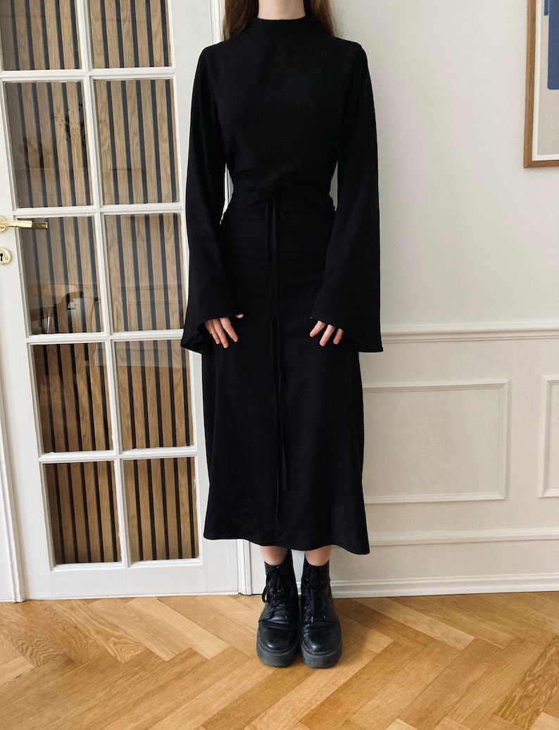 Puede incluir: Un vestido negro con mangas largas y acampanadas y cuello alto. El vestido tiene una cintura ce&ntilde;ida con cord&oacute;n y una falda midi. La persona lleva botas de combate negras.