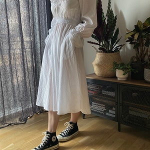 May include: A white, long-sleeved, ruffled dress with a cinched waist and pockets. The dress is paired with black high-top sneakers.
