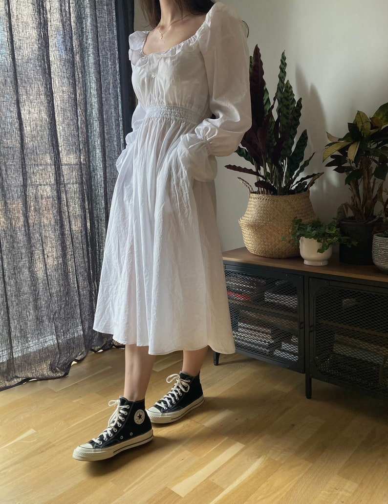 May include: A white, long-sleeved, ruffled dress with a cinched waist and pockets. The dress is paired with black high-top sneakers.