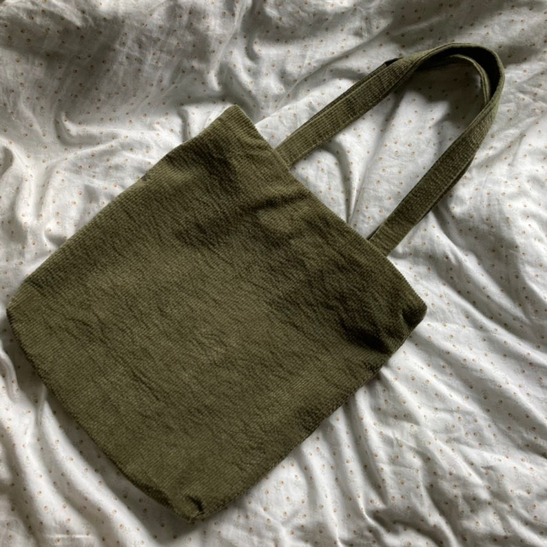 May include: A green corduroy tote bag with two long straps. The bag is lying on a white surface with a small pattern.
