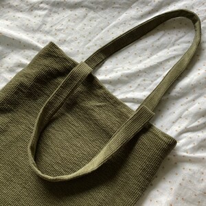 May include: A green corduroy tote bag with two long straps. The bag is made of a soft, textured fabric.