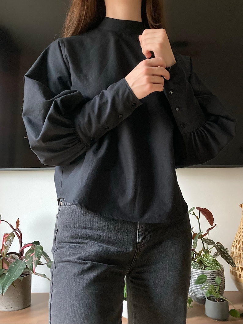 May include: A black mock neck shirt with long sleeves and buttoned cuffs. The shirt has a slightly puffed sleeve. The shirt is paired with dark wash jeans.