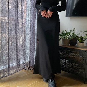 K&ouml;nnte beinhalten: Ein schwarzes, schulterfreies Maxikleid mit langen &Auml;rmeln. Das Kleid hat einen figurbetonten Oberteil und einen flie&szlig;enden Rock. Das Model tr&auml;gt schwarze Plateauschuhe.
