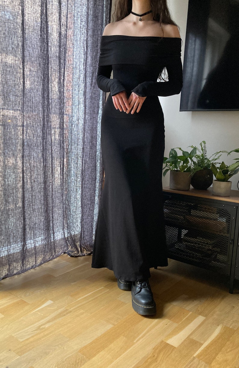 K&ouml;nnte beinhalten: Ein schwarzes, schulterfreies Maxikleid mit langen &Auml;rmeln. Das Kleid hat einen figurbetonten Oberteil und einen flie&szlig;enden Rock. Das Model tr&auml;gt schwarze Plateauschuhe.