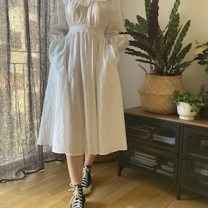 May include: A white, long-sleeved dress with a ruffled neckline and a cinched waist. The dress has a flowing skirt and pockets on the sides. The model is wearing black high-top sneakers.