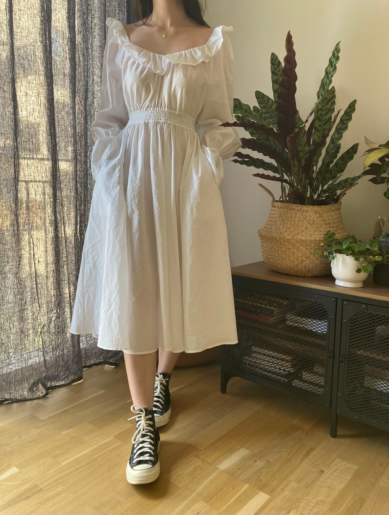 May include: A white, long-sleeved dress with a ruffled neckline and a cinched waist. The dress has a flowing skirt and pockets on the sides. The model is wearing black high-top sneakers.