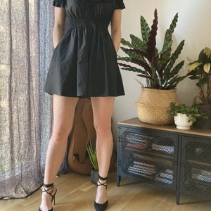May include: A black mini dress with a ruffled neckline and short sleeves. The dress has a fitted bodice and a flowy skirt. The model is wearing black heels with lace-up straps.