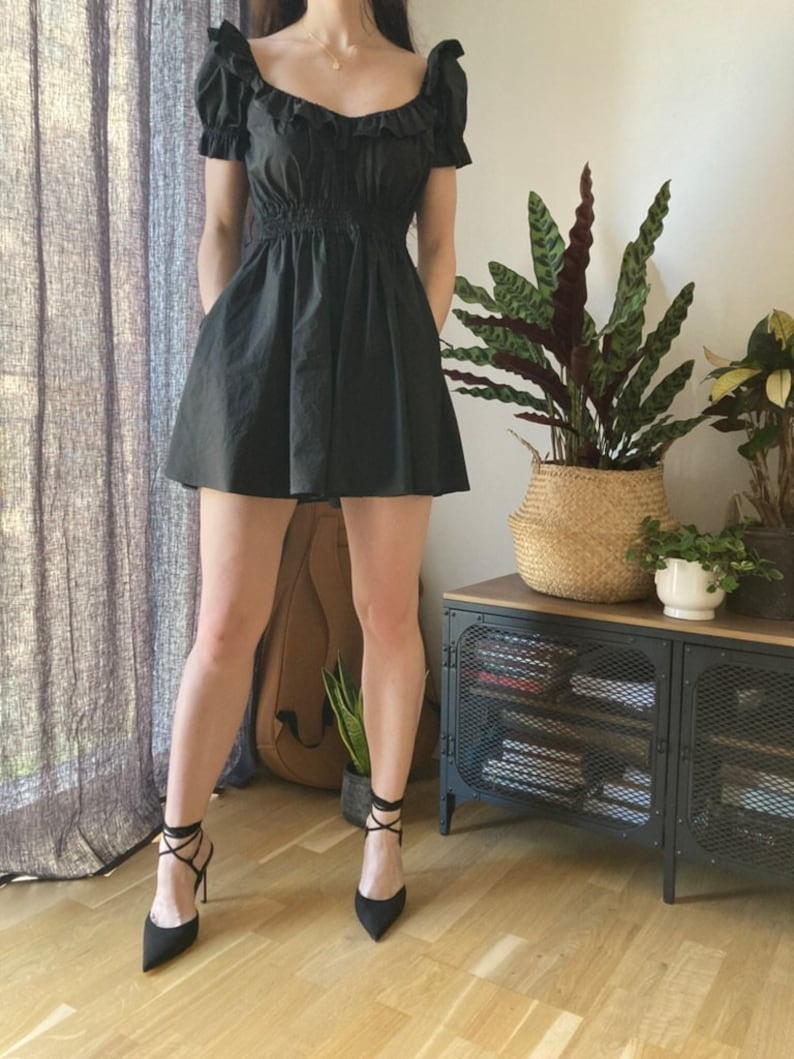 May include: A black mini dress with a ruffled neckline and short sleeves. The dress has a fitted bodice and a flowy skirt. The model is wearing black heels with lace-up straps.