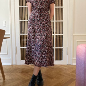 May include: A black midi dress with a floral print featuring red, white, and black flowers. The dress has a mock neck and short sleeves. The model is wearing black ankle boots.