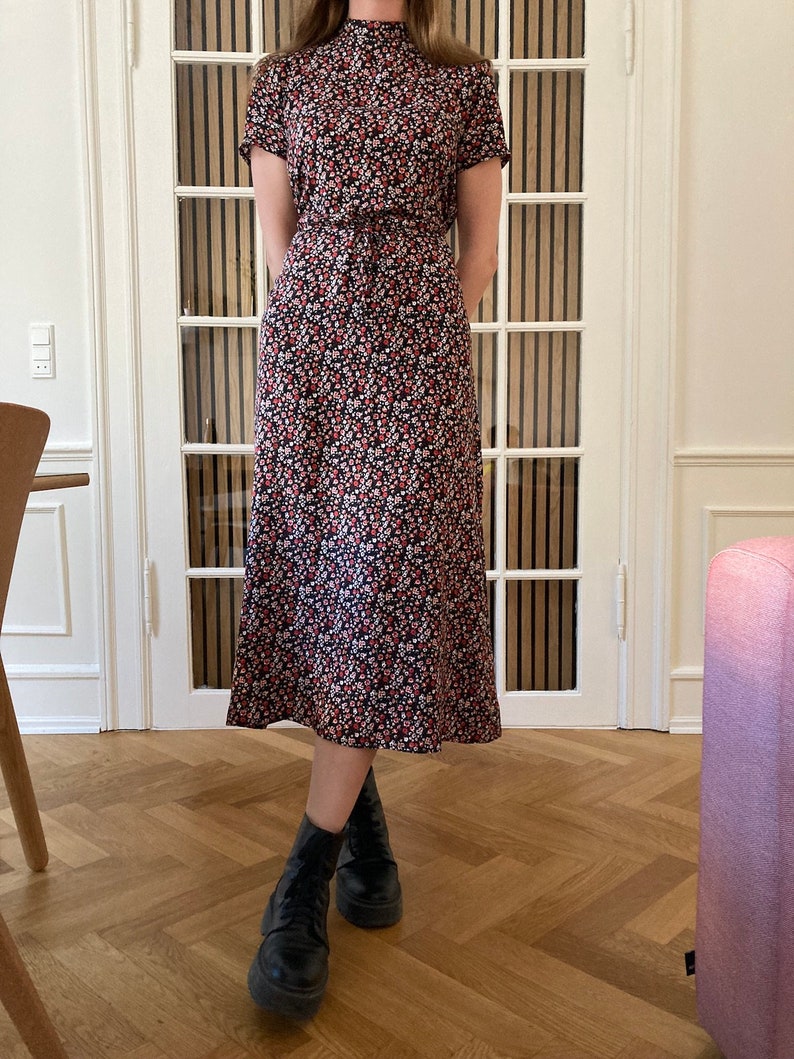 May include: A black midi dress with a floral print featuring red, white, and black flowers. The dress has a mock neck and short sleeves. The model is wearing black ankle boots.