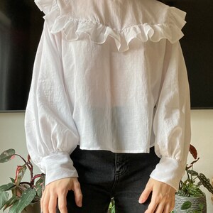 May include: A white, long-sleeved blouse with a ruffled neckline and puffed sleeves. The blouse is cropped and has a relaxed fit.