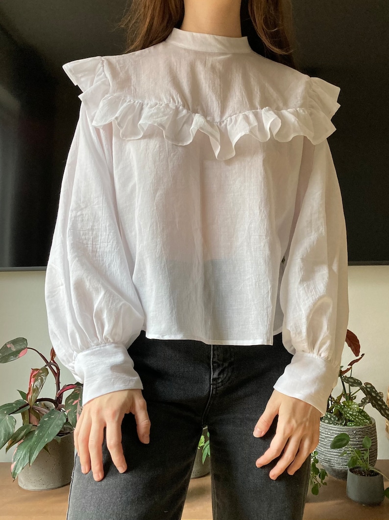 May include: A white, long-sleeved blouse with a ruffled neckline and puffed sleeves. The blouse is cropped and has a relaxed fit.