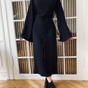 Puede incluir: Vestido negro con mangas largas y acampanadas y cuello alto. El vestido tiene un lazo en la cintura y se combina con botas negras. El vestido es de largo midi y est&aacute; hecho de una tela texturizada.