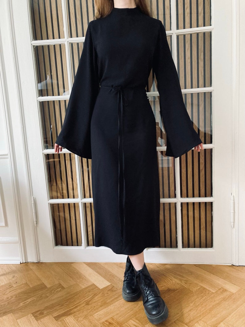 Puede incluir: Vestido negro con mangas largas y acampanadas y cuello alto. El vestido tiene un lazo en la cintura y se combina con botas negras. El vestido es de largo midi y est&aacute; hecho de una tela texturizada.