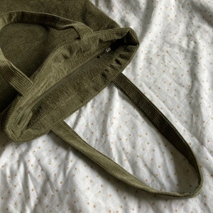 May include: A green corduroy tote bag with a zipper closure and two handles. The bag is lying on a white fabric with a floral pattern.