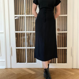 Puede incluir: Un vestido midi negro con mangas cortas y cuello alto. El vestido tiene una cintura ajustable con cord&oacute;n y se combina con botas negras con cordones. El fondo incluye una puerta blanca con paneles de madera verticales.