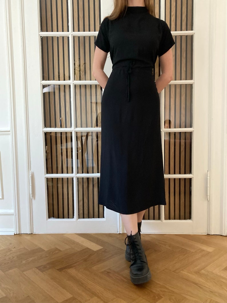 Puede incluir: Un vestido midi negro con mangas cortas y cuello alto. El vestido tiene una cintura ajustable con cord&oacute;n y se combina con botas negras con cordones. El fondo incluye una puerta blanca con paneles de madera verticales.