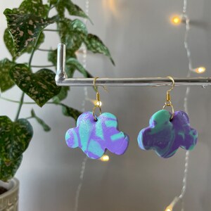 May include: A pair of cloud-shaped earrings with a marbled purple and green design. The earrings are made of polymer clay and have gold hooks.