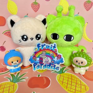 May include: A collection of plush toys and figurines, including a white cat-like plush, a green alien-like plush, and a blue bear figurine. A sticker with a rainbow and the words "Fruit Paradise" is also present. The background features a pink pattern with fruit.