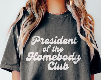 Può includere: Una maglietta nera con testo bianco che recita "President of the Homebody Club" in un font retrò.