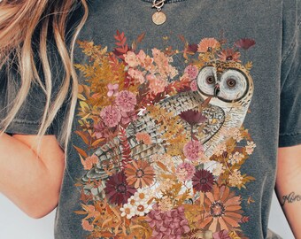 Könnte beinhalten: Ein graues T-Shirt mit einem grafischen Aufdruck eines Eulen, umgeben von Blumen in Braun-, Rosa- und Gelbtönen. Der Text "pepper" ist in der rechten unteren Ecke des Bildes sichtbar.
