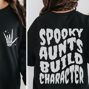 Pode incluir: Camisola de moletom preta com uma mão de esqueleto fazendo o sinal de "Eu te amo" na frente e o texto "Spooky Aunts Build Character" nas costas.