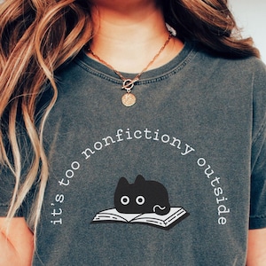 May include: Dark grey t-shirt featuring the text "it's too nonfictiony outside" encircling a graphic of a black cat on an open book. The shirt is a relaxed, casual style.