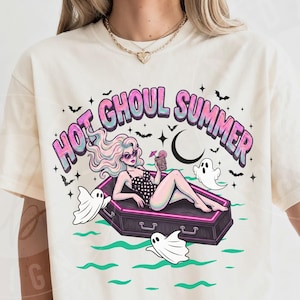 May include: Ivory-colored t-shirt with a graphic that reads "Hot Ghoul Summer." The design features a woman in a coffin, surrounded by ghosts and bats. The shirt is a relaxed fit.