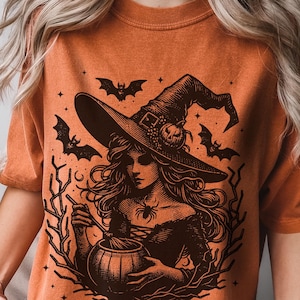 May include: Orange t-shirt with a black and white illustration of a witch stirring a cauldron. The witch is wearing a pointed hat and has long flowing hair. The illustration is surrounded by bats and stars.