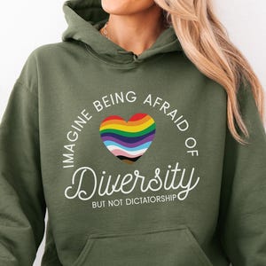 May include: Military green hoodie with a rainbow heart graphic and the words "Imagine Being Afraid of Diversity But Not Dictatorship." The hoodie has a front pocket and a drawstring hood. The text "MILITARY GREEN" is at the bottom.