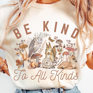 Pode incluir: Uma camiseta bege com um gráfico marrom de animais da floresta e cogumelos. O texto "Be Kind To All Kinds" é impresso em marrom na camiseta.