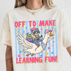 May include: Ivory-colored t-shirt with a colorful graphic of a goose carrying a raccoon and a frog, with the text "OFF TO MAKE LEARNING FUN!" in a playful font. The design includes stars and stripes.