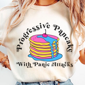 May include: A white t-shirt with a graphic of a stack of pancakes with blue and pink frosting and a cherry on top. The text "Progressive Pancake with Panic Attacks" is written around the graphic.