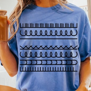 May include: A light blue t-shirt with a black graphic design featuring various line patterns. The shirt is labeled "MYSTIC BLUE" at the bottom right. The person is wearing white socks with black stripes.