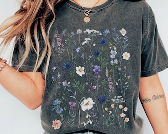 May include: A charcoal grey t-shirt with a floral print featuring various shades of blue, purple, white, and yellow flowers. The shirt has a crew neck and short sleeves.