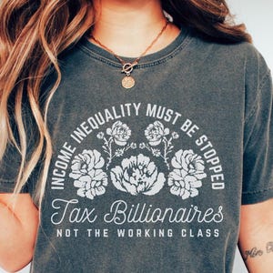 Può includere: Una maglietta grigia scura con una stampa grafica bianca che recita "Income Inequality Must Be Stopped Tax Billionaires Not The Working Class". La grafica è circondata da disegni floreali bianchi.