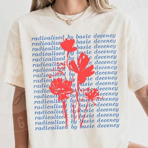 以下が含まれることがあります： アイボリーの半袖Tシャツで、radicalized by basic decencyという青いテキストが繰り返されるパターンがプリントされています。テキストの上には、鮮やかな赤で描かれた3つの様式化された花のイラストが重ねられています。Tシャツはクルーネックで、ゆったりとしたフィット感です。