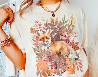 May include: A white t-shirt with a graphic of a fox surrounded by autumn leaves, mushrooms, and berries. The text "ivory" is written in the bottom right corner of the image.