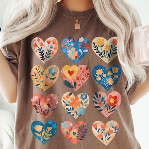 May include: Brown t-shirt with a floral heart design. The shirt has a relaxed fit and is made of soft material. The design features twelve heart shapes, each filled with unique floral patterns in various colours. The word "espresso" is at the bottom.