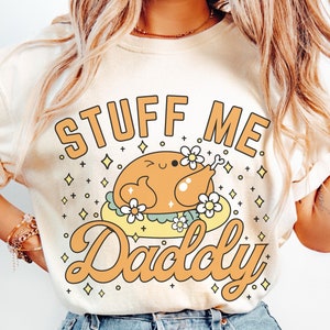 Puede incluir: Una camiseta blanca con un gráfico de un pavo de dibujos animados con el texto "Stuff Me Daddy" en una fuente retro.