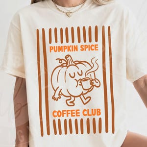 May include: An ivory short-sleeve t-shirt featuring a brown and orange graphic. The design includes vertical stripes, a cartoon pumpkin character holding a steaming mug, and the text PUMPKIN SPICE COFFEE CLUB printed in a bold, sans-serif font.