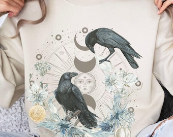 May include: A beige sweatshirt with a graphic design of two black ravens, a moon, and a sun with a floral design. The sweatshirt is worn by a person sitting on a couch.