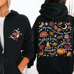 May include: Black zip-up hoodie with a design on the front and back. The front features a crescent moon, butterflies, and flowers. The back has the text "cozy blankets, crunching leaves, crackling fires, pumpkin everything" with fall-themed illustrations.