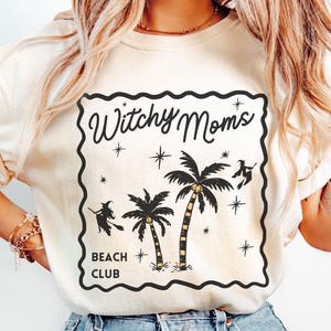 K&ouml;nnte beinhalten: Elfenbeinfarbenes T-Shirt mit einem schwarzen Aufdruck "Witchy Moms Beach Club". Das Design zeigt Palmen, Sterne und Hexen auf Besen. Das Shirt hat eine lockere Passform.