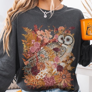 Pode incluir: Uma camisola de manga comprida cinzenta escura com uma estampa gr&aacute;fica de uma coruja cercada por flores de outono.