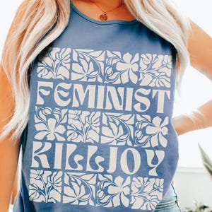May include: Blue jean tank top with the words "FEMINIST KILLJOY" in white block letters. The text is set against a white floral design. The tank top is a solid blue color.