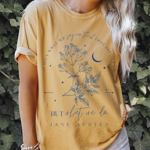以下が含まれることがあります： マスタードイエローのTシャツに、花柄のデザインと「私たちを定義するのは、私たちが言うことや考えることではなく、私たちがすることです。」というジェーン・オースティンの言葉が書かれています。