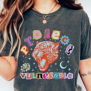May include: Dark gray t-shirt with a colorful graphic design. The design features a roaring tiger with the words "Protect the Vulnerable" in a mix of bright colors. The shirt has a vintage, worn look.